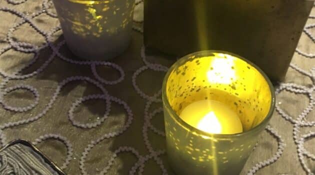 The flame of a candle burns. The candle is in a translucent container, set on a brocade tablecloth.