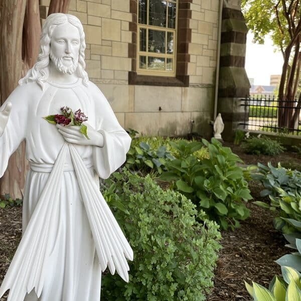 Statue of Jesus, the Divine Mercy, holding his hand up in blessing while positioned in a garden outside a chapel