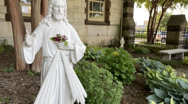 Statue of Jesus, the Divine Mercy, holding his hand up in blessing while positioned in a garden outside a chapel