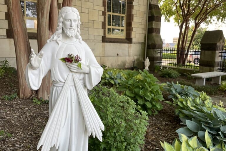 Statue of Jesus, the Divine Mercy, holding his hand up in blessing while positioned in a garden outside a chapel