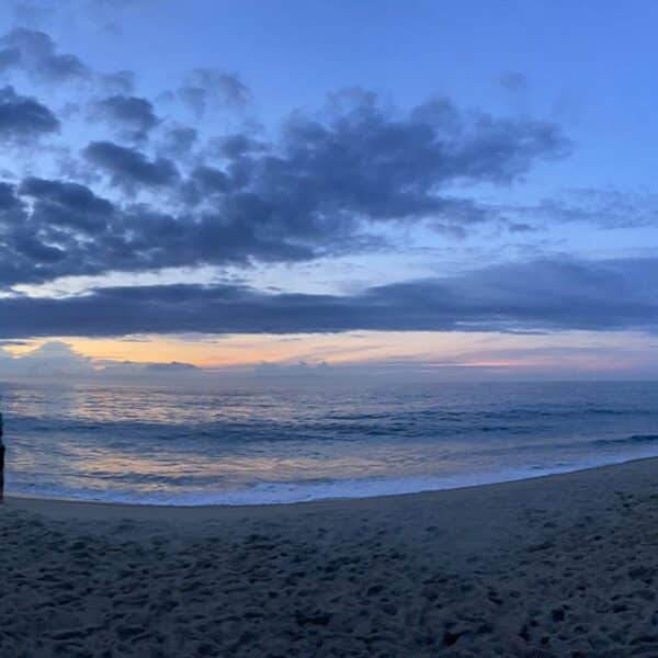 Wide shot of a sunrise on the beach, with a figure standing toward the left watching the light come into the sky