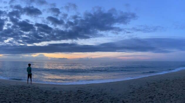Wide shot of a sunrise on the beach, with a figure standing toward the left watching the light come into the sky