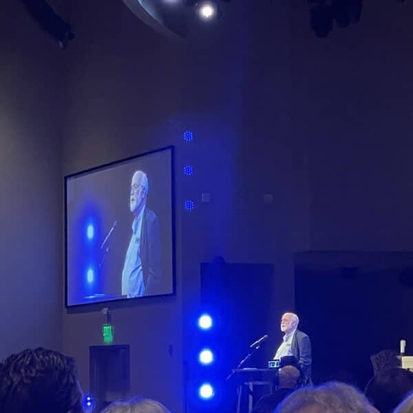 Fr. Greg Boyle, S.J., speaks from a podium with a crucifix hanging on one side and his image displayed on a video screen on the other side