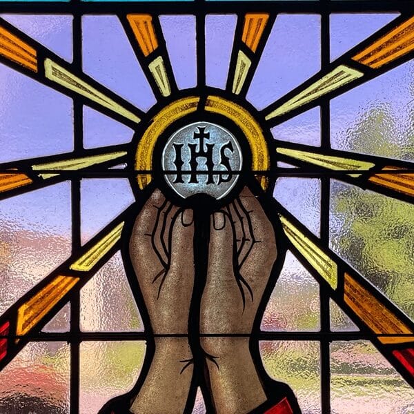 Stained glass window shows hands lifting a host with IHS on it